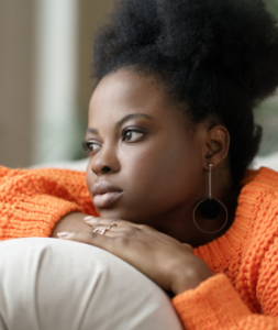 A Black woman in an orange sweater sitting on a couch deep in thought.